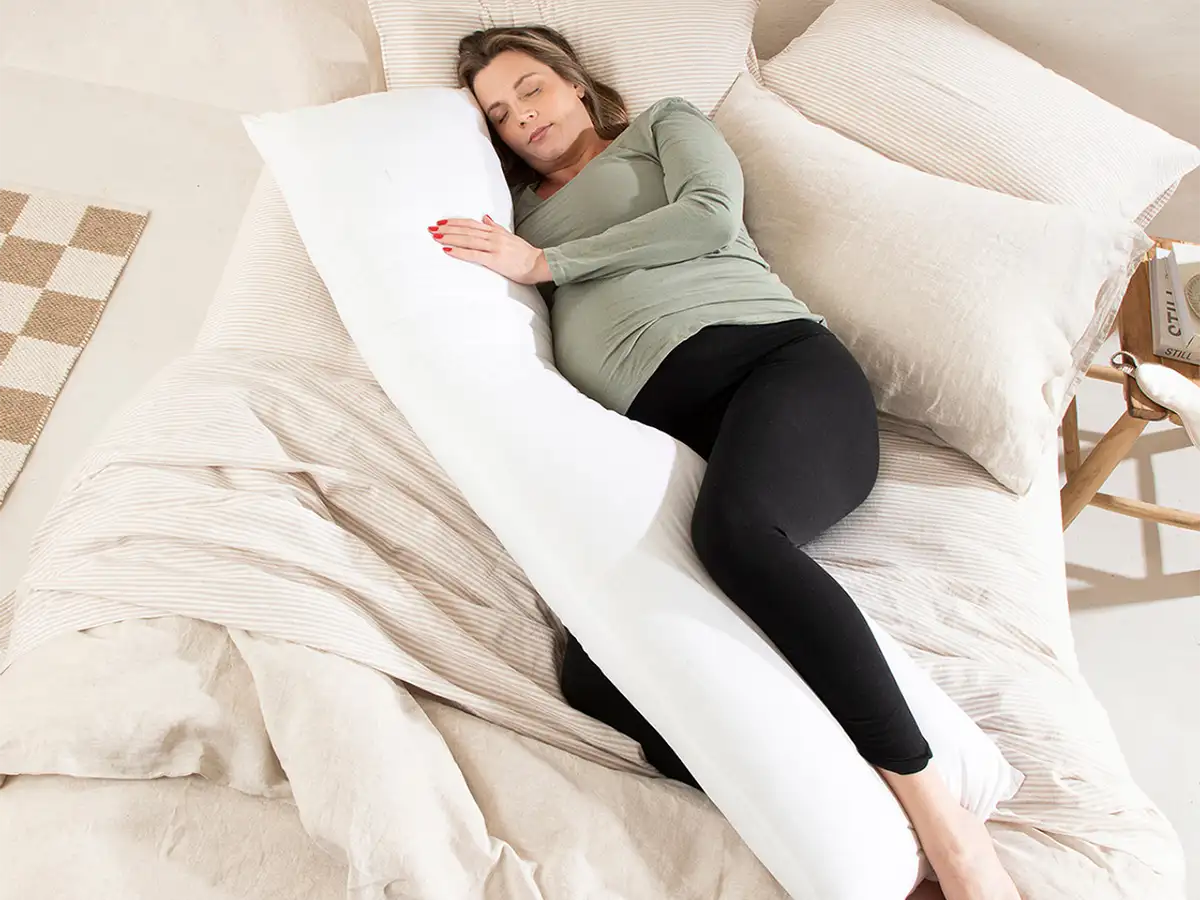 A pregnant woman comfortably rests on her side in bed, using the Comodo Original Body Pillow for support.